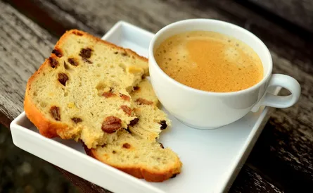 A slice of raisin bread on a plate next to cup of coffee